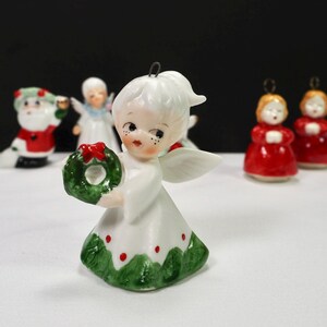 Puede incluir: Una figura de &aacute;ngel navide&ntilde;o de cer&aacute;mica con una corona verde y detalles rojos. El &aacute;ngel lleva un vestido blanco con adornos verdes y puntos rojos. Otras figuras navide&ntilde;as, incluyendo Pap&aacute; Noel y dos &aacute;ngeles con t&uacute;nicas rojas, est&aacute;n en el fondo.