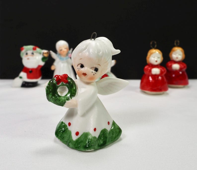 Puede incluir: Una figura de &aacute;ngel navide&ntilde;o de cer&aacute;mica con una corona verde y detalles rojos. El &aacute;ngel lleva un vestido blanco con adornos verdes y puntos rojos. Otras figuras navide&ntilde;as, incluyendo Pap&aacute; Noel y dos &aacute;ngeles con t&uacute;nicas rojas, est&aacute;n en el fondo.
