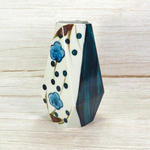 May include: A hexagonal ceramic vase featuring a cream-colored base with a dark teal vertical panel. The surface is decorated with hand-painted blue floral motifs and scattered dark blue dots, accented by thin brown brushstrokes on the cream side.