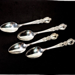 May include: Four silver spoons with ornate floral designs on the handles are arranged on a black surface. The spoons have a reflective, polished finish, and the handles feature detailed embellishments.