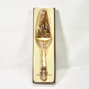 May include: A gold-colored cake server in its original packaging. The server is triangular with a long handle and is wrapped in clear plastic. The packaging has the brand name "HANSEAT Qualitats Bestecke" printed on it.