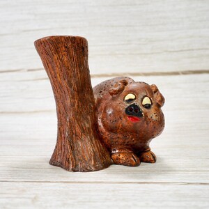 May include: A brown ceramic pig figurine with a tree trunk. The pig has a red mouth, black nose, and white eyes. The tree trunk is brown with textured details. The figurine is on a light wood surface.