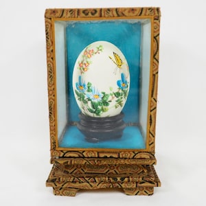 May include: A decorative egg with a painted floral and butterfly design, displayed in a glass case with a wooden frame and stand. The egg features blue and white flowers, and a yellow butterfly.