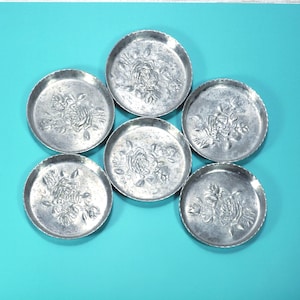May include: Six small, round, silver-colored metal coasters. Each coaster features a raised floral design in the center. The coasters are arranged on a turquoise surface, creating a decorative and functional set.