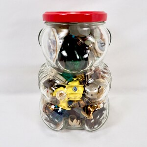May include: A clear glass bear-shaped jar with a red lid filled with a variety of buttons, trinkets, and small toys.