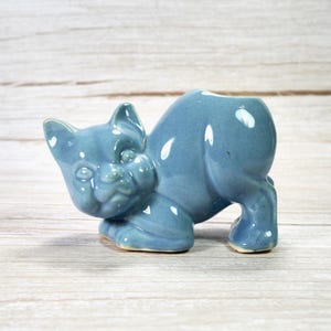 May include: A light blue ceramic cat figurine in a playful pose. The cat is on all fours, with its head turned to the side. The figurine has a glossy finish and is likely a planter or decorative item.