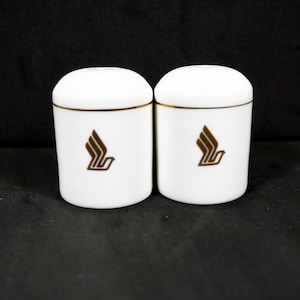 May include: Two white ceramic salt and pepper shakers with a gold trim and the Singapore Airlines logo in gold.