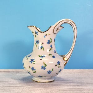 May include: A white porcelain pitcher featuring a delicate pattern of small blue cornflowers with green stems. The vessel has a curved handle, a scalloped rim, and a base, all accented with thin gold trim along the edges and handle details.