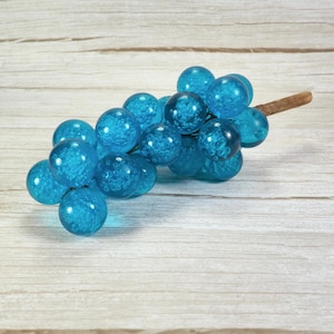 May include: A decorative cluster of translucent blue glass spheres arranged to resemble a bunch of grapes. Each sphere contains visible internal air bubbles and is attached to a short, thin, light brown wooden stem extending from one side.