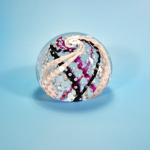 May include: A clear glass paperweight with a swirling design of white, black, and purple. The paperweight is round and has small bubbles throughout, resting on a light blue surface.
