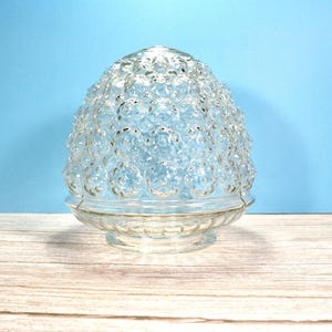 May include: A clear glass light fixture shade with a textured, bubble-like pattern. The shade has a rounded, egg-like shape and sits on a circular base. The background is a light blue.