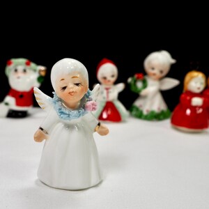 Puede incluir: Una colecci&oacute;n de peque&ntilde;as figuras navide&ntilde;as de cer&aacute;mica. El primer plano presenta un &aacute;ngel blanco con los ojos cerrados, un cuello azul y peque&ntilde;as alas. Otras figuras incluyen a Pap&aacute; Noel, &aacute;ngeles y figuras con vestidos rojos.