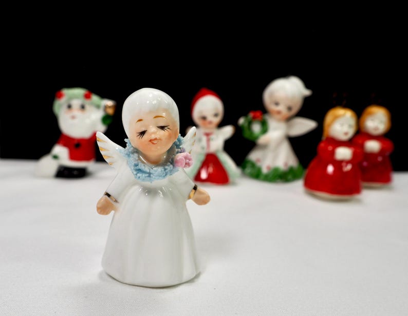 Puede incluir: Una colecci&oacute;n de peque&ntilde;as figuras navide&ntilde;as de cer&aacute;mica. El primer plano presenta un &aacute;ngel blanco con los ojos cerrados, un cuello azul y peque&ntilde;as alas. Otras figuras incluyen a Pap&aacute; Noel, &aacute;ngeles y figuras con vestidos rojos.