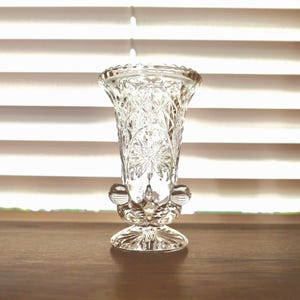 May include: A clear, vintage-style glass vase with intricate starburst and diamond patterns. The vase has a flared top, a decorative base, and two ornate handles. The vase is displayed on a wooden surface in front of a window with white blinds.