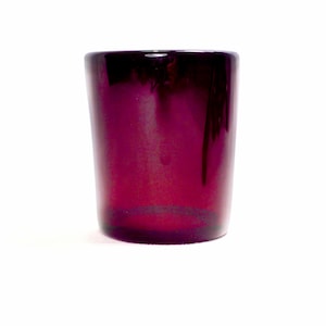 May include: A cylindrical, deep purple glass tumbler. The glass has a glossy finish and a slightly tapered shape, wider at the top. The color transitions from a darker shade at the top to a lighter hue towards the base.
