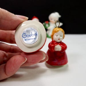 Puede incluir: Peque&ntilde;a figura Goebel roja y blanca de un &aacute;ngel rezando con cabello dorado y t&uacute;nica roja. La base de la figura tiene una pegatina plateada con el texto "Goebel W. Germany". Otras figuras son visibles en el fondo.