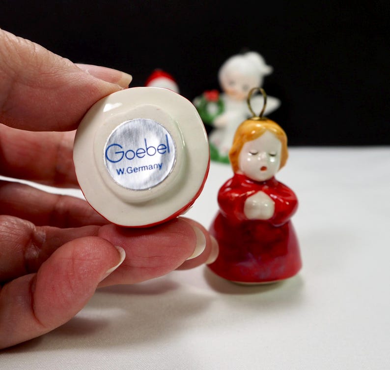 Puede incluir: Peque&ntilde;a figura Goebel roja y blanca de un &aacute;ngel rezando con cabello dorado y t&uacute;nica roja. La base de la figura tiene una pegatina plateada con el texto "Goebel W. Germany". Otras figuras son visibles en el fondo.