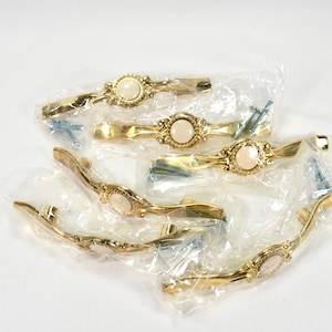 May include: A collection of six gold-toned cabinet pulls, each featuring an oval, off-white center stone. The pulls are packaged in clear plastic and include mounting screws. The design is ornate and vintage.