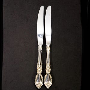 May include: Two silver-colored knives with ornate handles are displayed against a black background. The blades are smooth and reflective, while the handles feature a detailed, floral design. The knives are likely for formal dining.