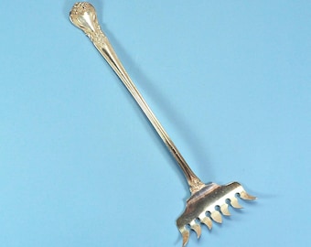 May include: A silver-colored spaghetti server with a long handle and a decorative floral design at the top. The serving end has multiple curved tines. The server is angled against a light blue background.