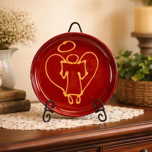 May include: A round, red decorative plate with a gold angel design, displayed on a black metal stand. The angel is inside a heart shape. The plate is on a wooden surface with a white lace doily.