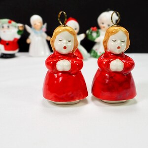 Puede incluir: Dos adornos navide&ntilde;os de cer&aacute;mica que representan a una ni&ntilde;a con un vestido rojo con las manos juntas. La ni&ntilde;a tiene el pelo rubio y los ojos cerrados. Un lazo dorado en la parte superior para colgar. Otras figuras navide&ntilde;as est&aacute;n en el fondo.