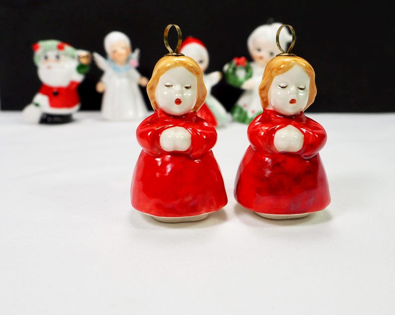 Puede incluir: Dos adornos navide&ntilde;os de cer&aacute;mica que representan a una ni&ntilde;a con un vestido rojo con las manos juntas. La ni&ntilde;a tiene el pelo rubio y los ojos cerrados. Un lazo dorado en la parte superior para colgar. Otras figuras navide&ntilde;as est&aacute;n en el fondo.