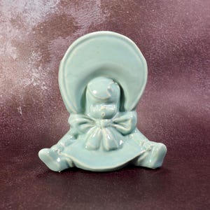 May include: A light blue ceramic figurine of a baby wearing a bonnet and a bow. The figurine is sitting with its legs extended. The baby's face is detailed with small features. The figurine is a vintage collectible.