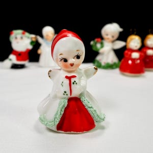 Puede incluir: Colecci&oacute;n de figuras navide&ntilde;as vintage. La figura central es una ni&ntilde;a con un vestido rojo y blanco con una gorra roja. Otras figuras incluyen a Pap&aacute; Noel, &aacute;ngeles y ni&ntilde;as con vestidos rojos. Las figuras son de cer&aacute;mica.