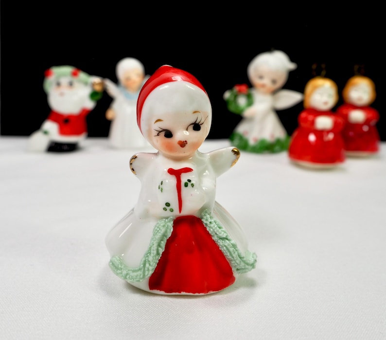 Puede incluir: Colecci&oacute;n de figuras navide&ntilde;as vintage. La figura central es una ni&ntilde;a con un vestido rojo y blanco con una gorra roja. Otras figuras incluyen a Pap&aacute; Noel, &aacute;ngeles y ni&ntilde;as con vestidos rojos. Las figuras son de cer&aacute;mica.