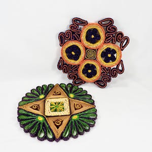 May include: Two woven pot holders, one with a starburst design in green, yellow, and brown, and the other with a circular design with four smaller circles in orange, yellow, and purple.