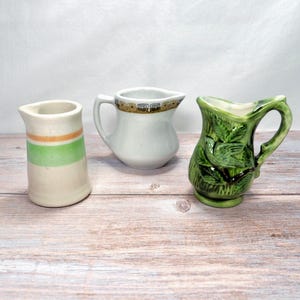 May include: Three small ceramic pitchers. One is white with green and orange stripes, another is white with a brown and gold rim, and the third is green with a leaf design.