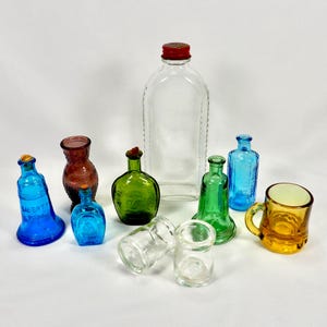 May include: A collection of colorful, vintage glass bottles and containers. Includes a clear, rectangular bottle with a red cap, a blue bell-shaped bottle with text, and a small amber mug.