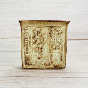 May include: A square, ceramic planter with a light green glaze and a brown rim. The planter features a raised, textured design with floral and geometric patterns. The design is divided into rectangular sections.