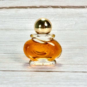 May include: A small, vintage perfume bottle with a rounded, amber-colored glass body and a gold-colored spherical cap. The bottle has a clear glass neck and sits on a wooden surface.