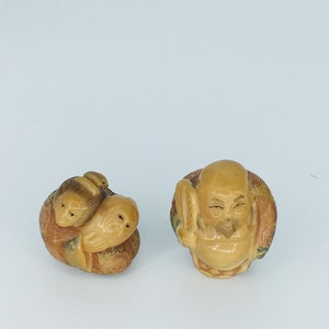 Pode incluir: Duas figuras esculpidas em cor de marfim. Uma retrata uma figura sentada com um leque, a outra mostra um grupo de figuras encolhidas. As esculturas têm traços faciais detalhados sobre um fundo branco.
