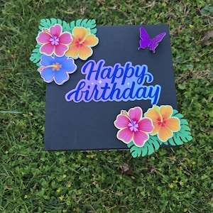 May include: A black card with the words "Happy birthday" written in blue and purple script. The card is decorated with colorful paper flowers and a purple glitter butterfly.