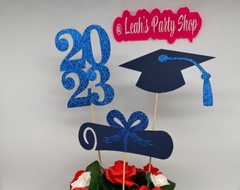 Puede incluir: Un centro de mesa de graduación con una caja de madera llena de rosas rojas y blancas y hojas verdes. Los recortes de purpurina azul incluyen el año "2023", un birrete y un diploma. El centro de mesa tiene el texto "@ Leah's Party Shop".