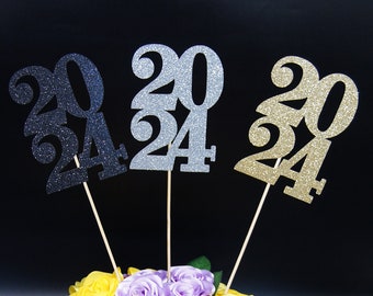 May include: A centerpiece featuring a burlap box with a white bow and three glitter-covered "2024" signs in black, silver, and gold, surrounded by yellow and purple silk roses.