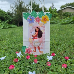 May include: White paper gift bag featuring a cartoon image of a Polynesian woman holding a pig. The bag is decorated with colorful paper flowers and green leaves. Pink and white flowers are scattered around the base of the bag.