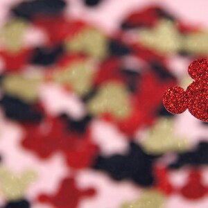 May include: A red glitter Mickey Mouse head cutout with a single ear on the left side, against a blurred background of red, gold, and black glitter shapes.