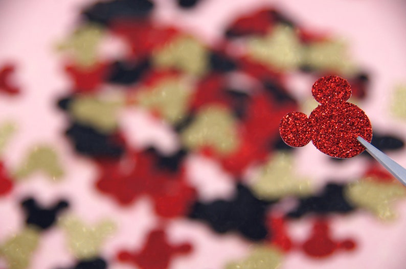 May include: A red glitter Mickey Mouse head cutout with a single ear on the left side, against a blurred background of red, gold, and black glitter shapes.