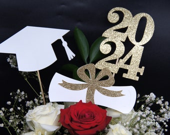 Puede incluir: Centro de mesa de graduación con rosas blancas, una rosa roja y aliento de bebé. El centro de mesa presenta un gorro de graduación blanco, un diploma blanco con un lazo de brillo dorado y brillo dorado 2024.
