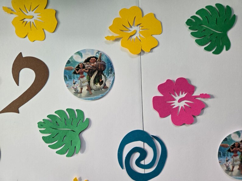 Pode incluir: Um conjunto de decora&ccedil;&otilde;es para festas com tema Moana, incluindo folhas verdes e marrons, flores de hibisco rosa e amarelas, um design de espiral azul e duas imagens redondas de Moana e Maui.