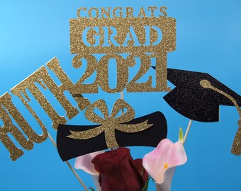 May include: Gold glitter graduation cake topper with the words "Congrats Grad 2021" and the name "Ruth". The topper also includes a black graduation cap, a diploma, and a bow.