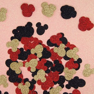 May include: A scattering of red, gold, and black glitter Mickey Mouse head confetti on a pink background.