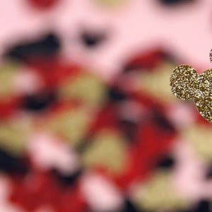 May include: A gold glitter Mickey Mouse head cutout against a blurred background of red, black, and pink.