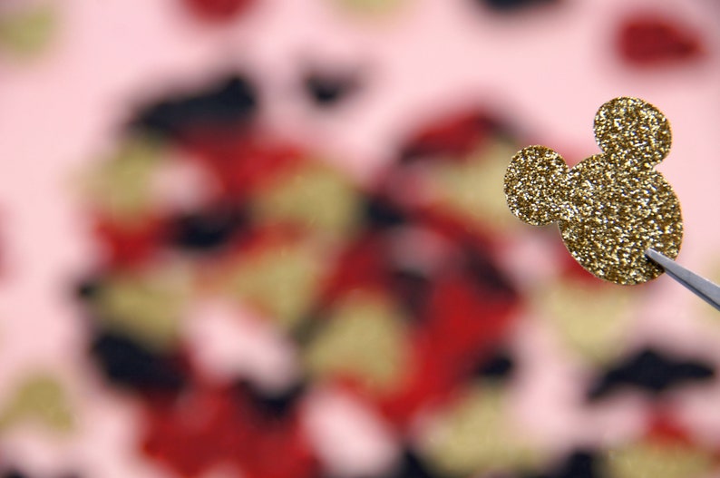 May include: A gold glitter Mickey Mouse head cutout against a blurred background of red, black, and pink.