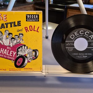 May include: A vintage vinyl record and album cover for "Shake, Rattle and Roll" by Bill Haley and his Comets. The album cover is yellow with a red hot rod illustration and the band's name. The record is black with a white label.
