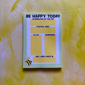 May include: A green notepad with the text "BE HAPPY TODAY" and "AFFIRMATION OF THE DAY". The notepad has sections for "POSITIVE VIBES", "TO DO", "REMINDERS", and "SELF CARE CHECK IN". A cartoon bee is in the bottom left corner.